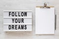 `Follow your dreams` words on a modern board, clipboard with blank sheet of paper on a white wooden surface, top view. Overhead,