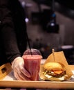 A tray with a burger on a foodcat. The chef& x27;s hand in a glove puts a glass with a drink on a tray. selective focus