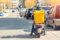Food delivery courier with big yellow backpack riding scooter on city street with traffic. Fast lunch takeaway delivery. Teenager
