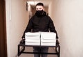 Food delivery man in black uniform, black protective mask and gloves delivers food to the house in boxes