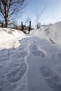 A footpath cleared of snow with footprints leads to an old rickety wicket and a fence among snowdrifts, against a blue sky on a Royalty Free Stock Photo