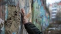 In the foreground a person stands with back to the camera hand pressed against a cracked and graffitied wall a stark