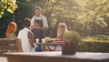 In a forest scenery, a very attractive waiter with a beard and hair tied back is taking orders from a table with three Royalty Free Stock Photo