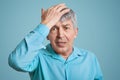 Forgetful handsome elederly wrinkled man in blue shirt, keeps hand on head, feels desperately as forgets about important meeting, Royalty Free Stock Photo