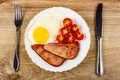 Fork, fried egg, slices of gammon and halves of tomato in plate, knife on wooden table. Top view Royalty Free Stock Photo