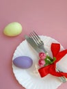 Fork, knife with red bowknot, white plate, colored eggs on pink background. Royalty Free Stock Photo