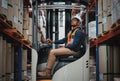 Happy forklift driver focused on carefully transporting stock from shelves around the floor of a large warehouse wearing