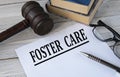 FOSTER CARE - words on white paper with the background of the judge`s hammer, glasses and pen