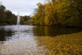 Fountain in fall nature with trees and lake with yellow fallen leaves in water Royalty Free Stock Photo