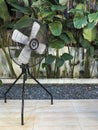 A four-bladed steel industrial fan mounted on a tripod at an outdoor al fresco garden themed cafe or restaurant