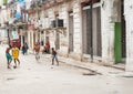 Four boys play football in urban street as people walk by surrounded by grungy old buildings neglected over years of US embargo