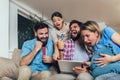 Four casual happy friends laughing shopping online together in a tablet sitting on a sofa in the living room