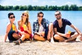Four friends sitting on lake beach with cocktails