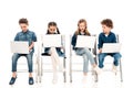 Four kids in denim clothes sitting on chairs and using laptops on white.