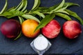 Four large peaches, a close-up of a peach sprig and a jar of cream on a black slate board.