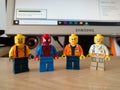 Four Lego toys, a spiderman and workers, on a computer desk in the room, and a lit monitor in the background