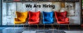 Four modern chairs against a grey wall with the words We Are Hiring written on it, signifying job opportunities and