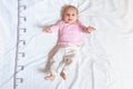 A four month happy baby in pink white clothes lying on a bed on which a measuring ruler for growth is drawn