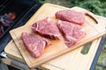 Four raw beef steaks lie on a wooden cutting board next to the grill