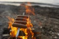 Four sausages on a metal skewer are roasting on an open fire at dusk against the sea