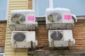 Four white air conditioning part of a split system hanging on a brick wall of a building covered in snow and icicles in winter in