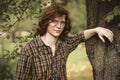 Foxy man with long carroty hair stands leaning on a tree trunk. Young handsome freckled guy in chequered shirt looking Royalty Free Stock Photo