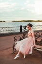 a fragile ballerina in a long pink dress with a full organza skirt sits on a wrought-iron wooden bench on the river bank