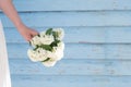 A fragile female hand is holding a small bouquet of white flowers against a blue plank wall. Use for design for flower shops,