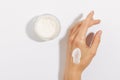 Fragile female hand with smear of cream and glass jar of facial moisturizer on white isolated background. Concept of skin care