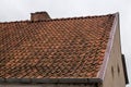 Fragment of old, worn brown tile roof with brick chimney, beige-plastered gable with small windows on it and a metal storm drain