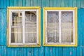 Fragment of wall of old wooden house with two windows with cracked paint on frames and holes between boards. Cornice with curtain Royalty Free Stock Photo