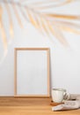 A frame with a blank canvas against a light wall and on a wooden tabletop with blurred palm leaf and cup of tea