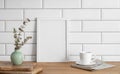 A frame with a blank canvas against a white tile wall and on a wooden tabletop with vase and cup of tea