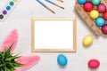 A frame, bunny ears, a flower pot with artificial green grass, colorful eggs in a cardboard box, paints and brushes. Royalty Free Stock Photo