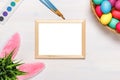 A frame, bunny ears, a flower pot with artificial green grass, colorful eggs in a cardboard box, paints and brushes. Royalty Free Stock Photo