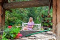 Framing view on a young blonde girl in a pink dress sits on a white wooden bench in the garden around flowers and plants with Royalty Free Stock Photo