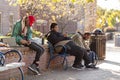 A group of poor people living on streets are sitting on a bench with their goods in backpacks.