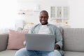 Free time. Happy middle-aged black man using laptop while resting on sofa at home Royalty Free Stock Photo