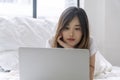 Freelance asian woman looking laptop lying down on white bed working at home. Teenager young girl using notebook learning online Royalty Free Stock Photo