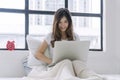 Freelance asian woman looking laptop sitting pillows on white bed working at home. Teenager young girl using notebook learning Royalty Free Stock Photo