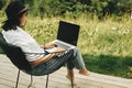 Freelance. Hipster girl with laptop sitting on wooden porch with beautiful view on woods and mountains. Stylish Young woman in hat