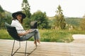 Freelance. Hipster girl with laptop sitting on wooden porch with beautiful view on woods and mountains. Stylish Young woman in hat