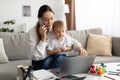 Freelance job and motherhood. Busy young mother working on laptop with toddler son on her lap, sitting on sofa at home Royalty Free Stock Photo