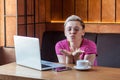 Freelancer with blonde short hair, in pink t-shirt and eyeglasses is sitting alone in cafe and working on laptop and sending air Royalty Free Stock Photo