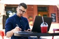 Freelancer man hand writing on paper notebook near his laptop in outdoor cafe. Young businessman in glasses taking notes Royalty Free Stock Photo