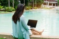 Freelancer using laptop working remotely near swimming pool. Young Asian traveler woman working on computer during her holiday Royalty Free Stock Photo