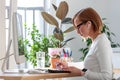 Freelancer woman taking notes with a pen, working on computer, using calculator to calculate the invoice, surrounded by plants. Royalty Free Stock Photo