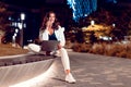 Freelancer woman talking on cellphone and using laptop in city park at night with neon lights on background, free space Royalty Free Stock Photo