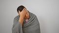 Freezing young latin man wrapped in cozy blanket, braving winter cold indoors, comfort against white isolated background, possibly