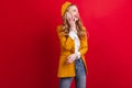 French woman in yellow jacket talking on phone. Studio shot of blonde girl in beret isolated on red background